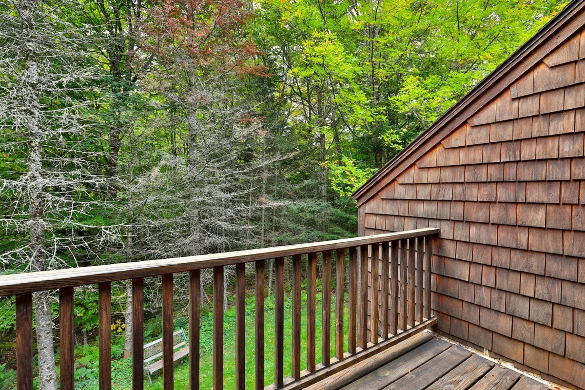 A peaceful exterior view featuring a wooden railing, shingle siding, and lush green surroundings. The space looks ideal for enjoying a morning coffee, reading a book, or simply soaking up the natural beauty, with hints of the changing seasons visible in the foliage.