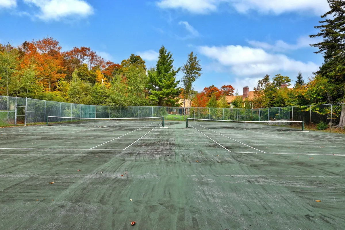 A spacious tennis court set amidst tall trees with a distant building visible, evoking an active yet relaxed atmosphere. The changing leaves suggest seasonal enjoyment and outdoor recreation.