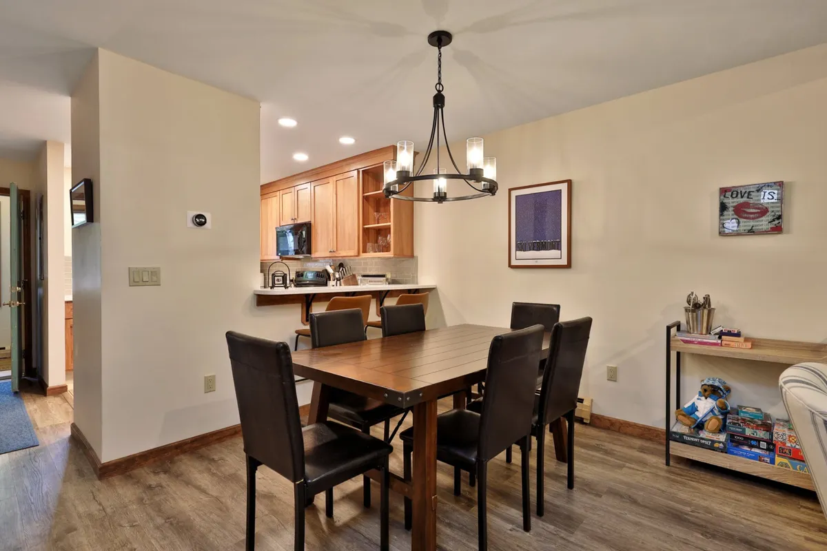 A spacious dining area adjacent to the kitchen, with a large table under a statement chandelier, shelves filled with games, and a kitchen visible in the background. The setup encourages gathering, shared meals, and family fun—key elements described in the section.