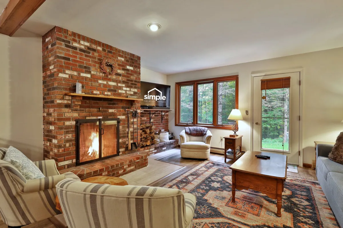 The image showcases a cozy living area centered around a glowing wood fireplace, with plush chairs arranged for relaxation, a rustic wooden table perfect for gatherings, and expansive windows letting in warm natural light. The inviting setup suggests comfort and togetherness, ideal for unwinding after a day on the slopes.