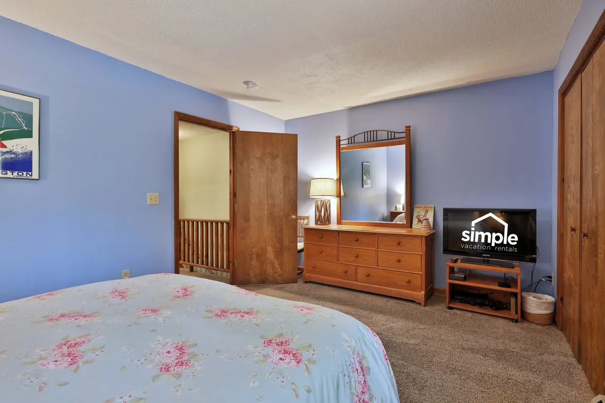 A bedroom featuring a plush bed with floral bedding, a wooden dresser, a mirror, and a television on top of the dresser. The space is tidy and inviting, with soft lighting and a window that brings in natural light. The TV is positioned for comfortable viewing from bed, and the room's decor is both cozy and refined.