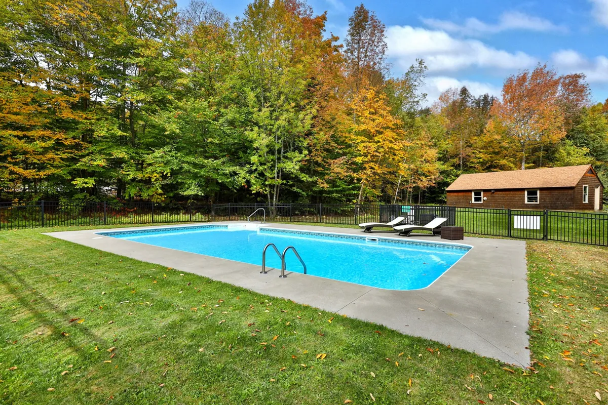 This image features an inviting outdoor swimming pool surrounded by lounge chairs and mature trees, with a small cabin in the background. The scene radiates a relaxed, summer atmosphere ideal for swimming, sunbathing, or gathering outdoors.