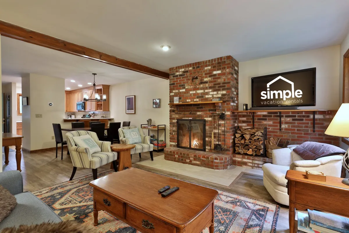 A welcoming living area featuring a plush seating arrangement centered around a wood-burning fireplace. The space includes a coffee table and a large smart TV, with warm ambient lighting creating an inviting mood. The setup is ideal for relaxing with family or friends, enjoying a cozy evening, or gathering for movie nights.