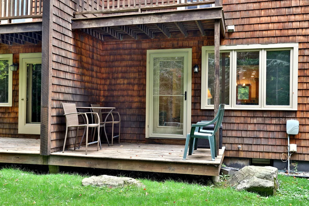 A private patio with a wooden deck and two comfortable chairs, overlooking a well-kept grassy area and bordered by stone accents. The outdoor setting is tranquil and inviting, ideal for morning coffee or relaxing with a drink while enjoying garden views and fresh air.