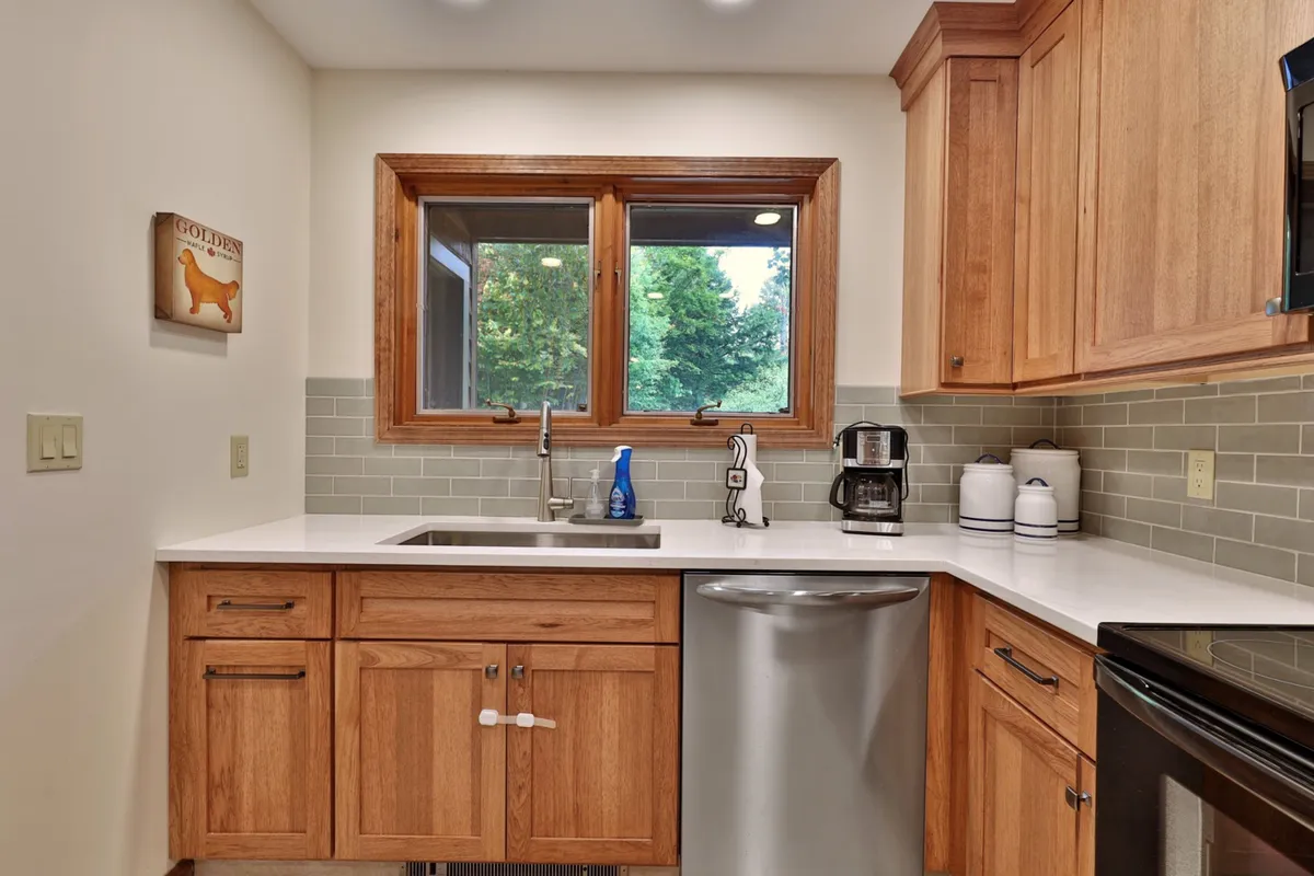 A cozy kitchen view featuring a sink positioned beneath a sunlit window, surrounded by wood cabinets and a convenient coffee maker on the countertop. The space exudes warmth and practicality, making it ideal for both casual mornings and more elaborate cooking sessions.