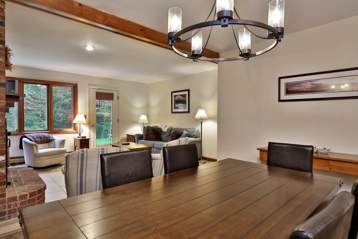 An open dining area featuring a spacious table with seating for multiple guests, illuminated by an elegant chandelier. Large windows allow natural light to fill the space, and the adjacent seating area further enhances the sense of connectivity and flow between spaces.